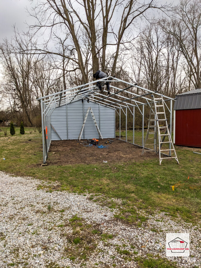 Front of metal garage currently being installed. It has one end fully enclosed, a vertical roof still being installed, two horizontal sides currently being installed, and one open end. A contractor is on the incomplete roof and there are tools and ladders scattered about the job site. This photo is just one example of the various types of pre-engineered metal buildings (PEMBs) we install.