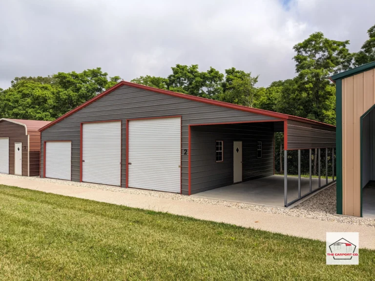 Corner of large metal garage by The Carport Co. that has a center section and two lean tos. The left lean to is fully enclosed and has a small 6x6 roll up door for storing lawn equipment and using it as other storage. The right lean-to is open, but does have a 3’ panel on the right side. The center section has two 10x10 roll up doors on the front end. The entire structure has dark gray horizontal siding and red trim.