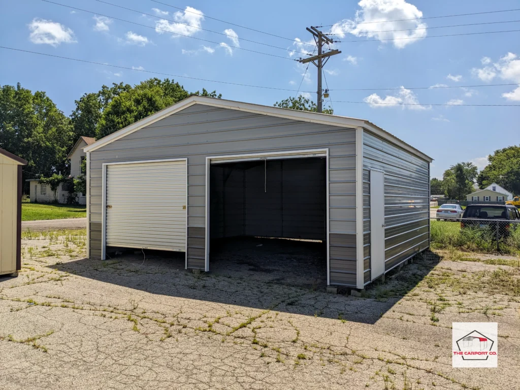 Blue steel garage with very dark gray wainscoting AKA two-tone addition, and white trim. The metal garage has horizontal siding, two 8x7 roll up doors on the front end, and a man door on the side. This is a standard configuration of a two car metal garage and one of The Carport Co.'s highest selling structures.